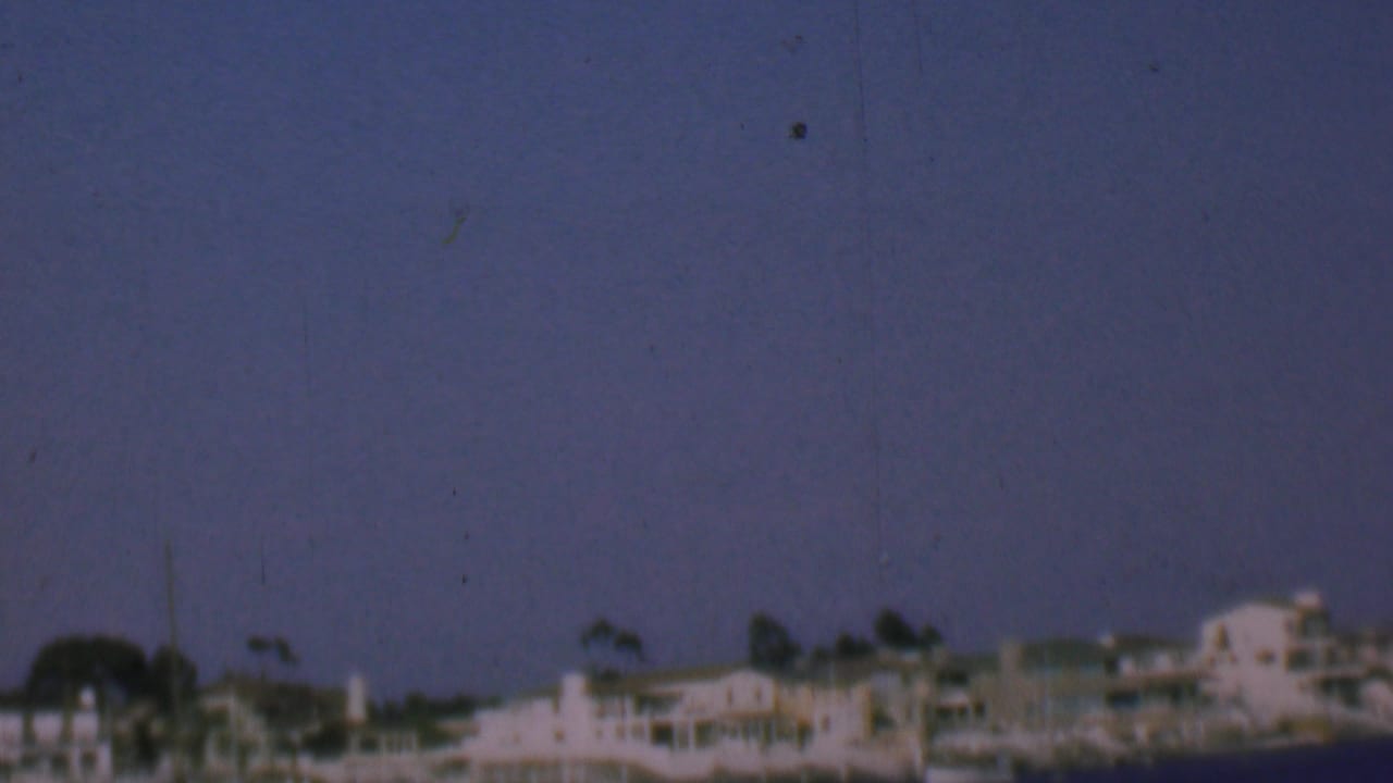 Soft 1961 8mm view of San Diego waterfront residences and palms seen from across the water, with the shoreline appearing slightly blurred.
