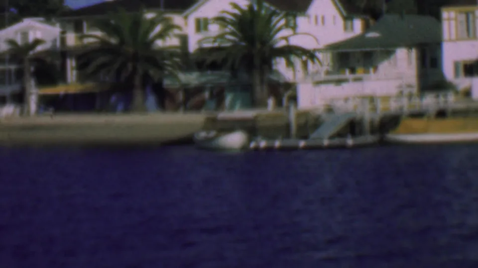 Soft 8mm frame of San Diego waterfront homes with a prominent palm, dock structures, small boats, and deep blue water in the foreground.