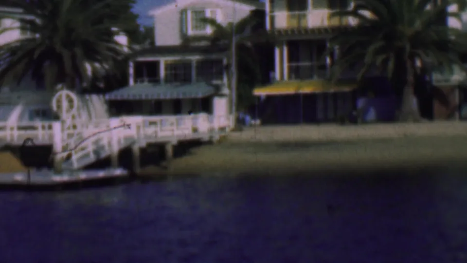 1961 San Diego waterfront frame with a white dock structure, palm trees, lawns, and residential houses along the shore.