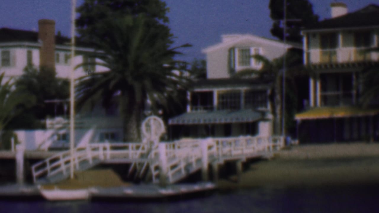 1961 San Diego home-movie frame showing white waterfront residences, palm trees, a white dock with railings, and a small boat along calm water.