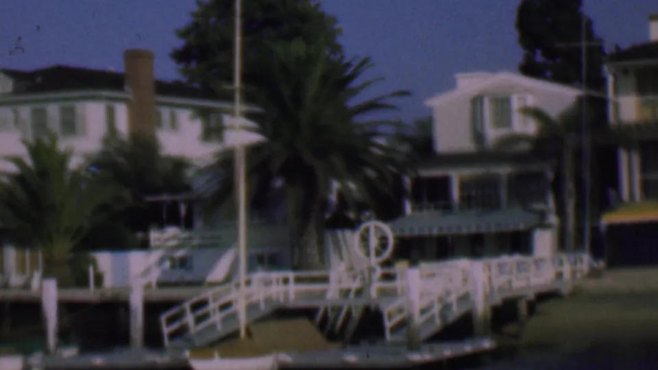1961 San Diego frame of a waterfront property with white residential buildings, palms, a dock structure, chimney, and mounted life preserver.
