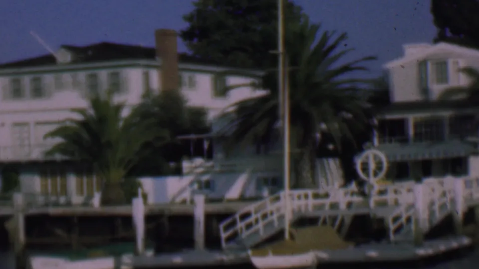 1961 San Diego home-movie view of white waterfront residences, a palm tree, dock railings, water, and a vertical pole in the foreground.