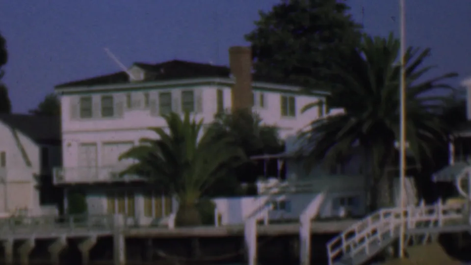 1961 San Diego frame showing a white waterfront residence with a prominent chimney, palms, dock posts, railings, and calm water in the foreground.