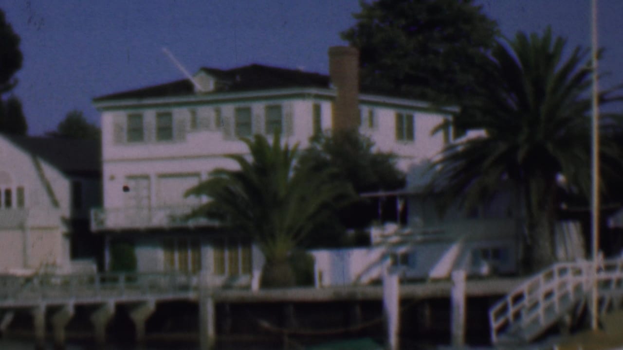 1961 San Diego 8mm frame showing a white waterfront house with green trim, a tall chimney, palm trees, dock railings, and a smaller white building at left.