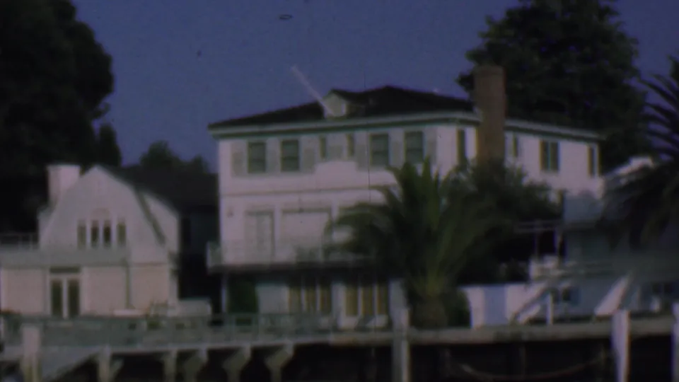 1961 San Diego frame with a white waterfront house, green window trim, tall chimney, palm fronds, dock posts, and a small gabled building beside it.