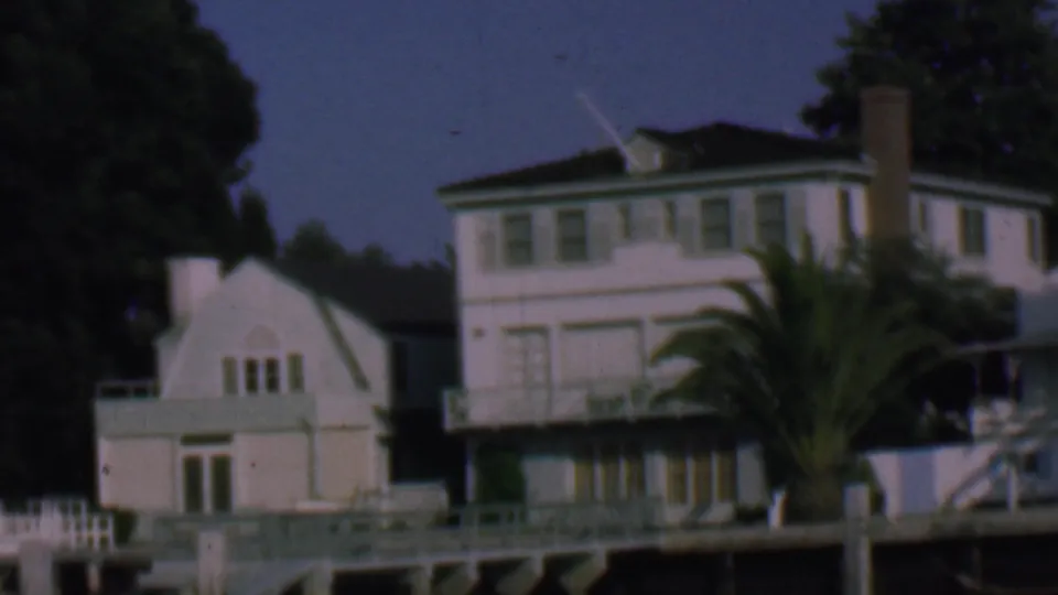 1961 San Diego 8mm view of a white house and smaller waterfront building with dock railings, pilings, a palm tree, and dark background trees.