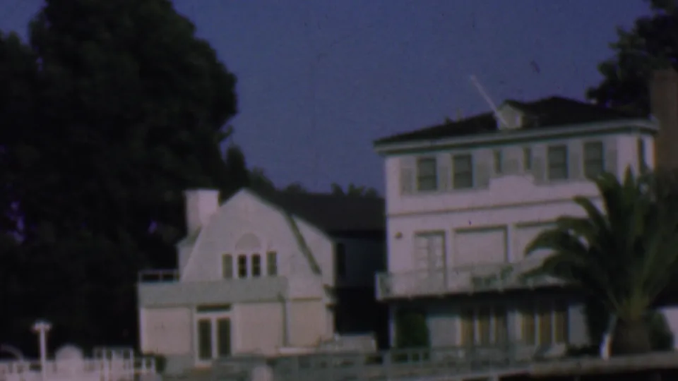 1961 San Diego frame showing the white waterfront residence, a smaller gabled building, palm fronds, dock railings, and dark trees in the background.