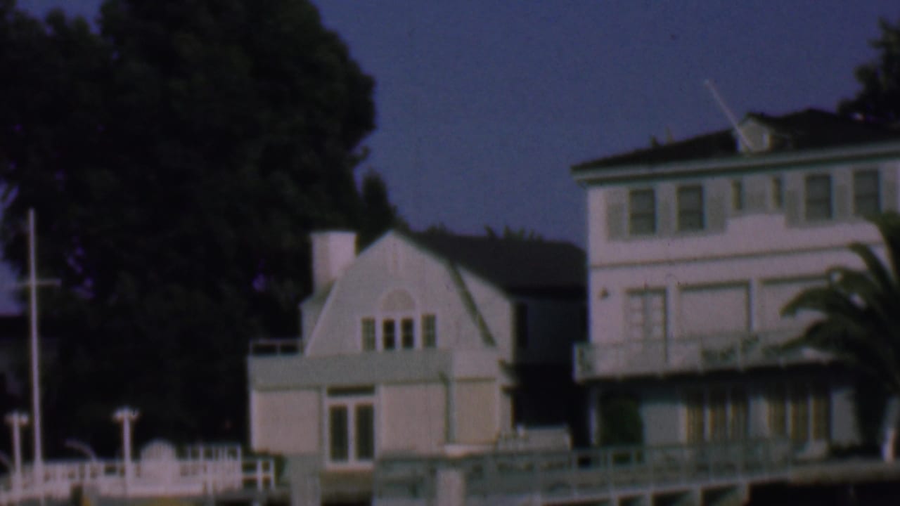 1961 San Diego-area 8mm frame showing white waterfront residences, including a small gambrel-roofed building, with mature trees behind them at dusk.