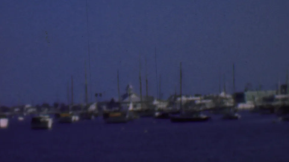 Soft 1961 home-movie frame of San Diego harbor with small boats in the foreground, dense masts, and low buildings along the distant shoreline.