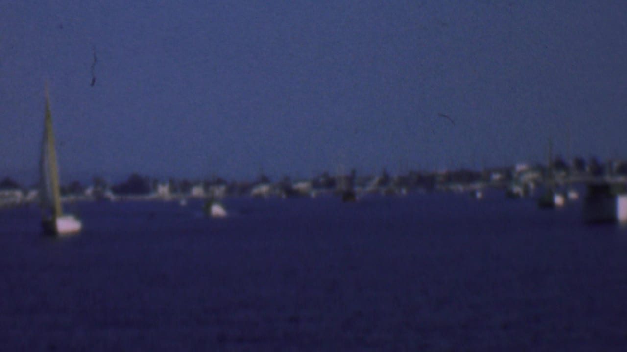 1961 San Diego home-movie frame showing anchored sailboats and motorboats on dark-blue bay water with a soft distant shoreline under hazy sky.