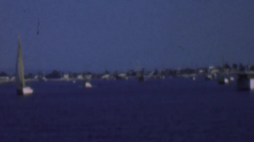 1961 San Diego home-movie frame showing anchored sailboats and motorboats on dark-blue bay water with a soft distant shoreline under hazy sky.