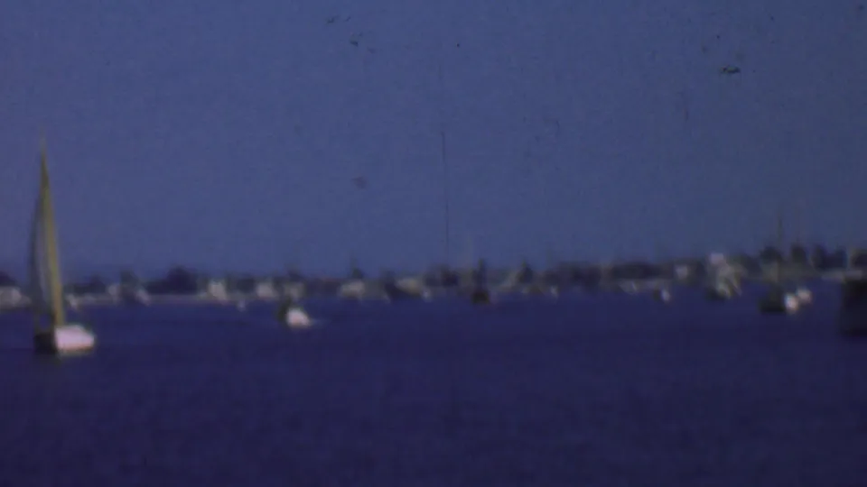 1961 San Diego home-movie frame with a sailboat partly at the left edge, calm water, anchored boats, and a low distant shore.