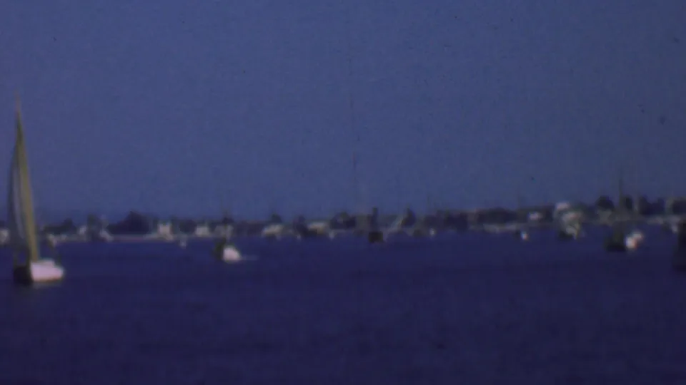 1961 San Diego bay frame showing a centered sailboat, scattered anchored vessels, deep-blue water, and a distant tree-lined shoreline in soft 8mm focus.