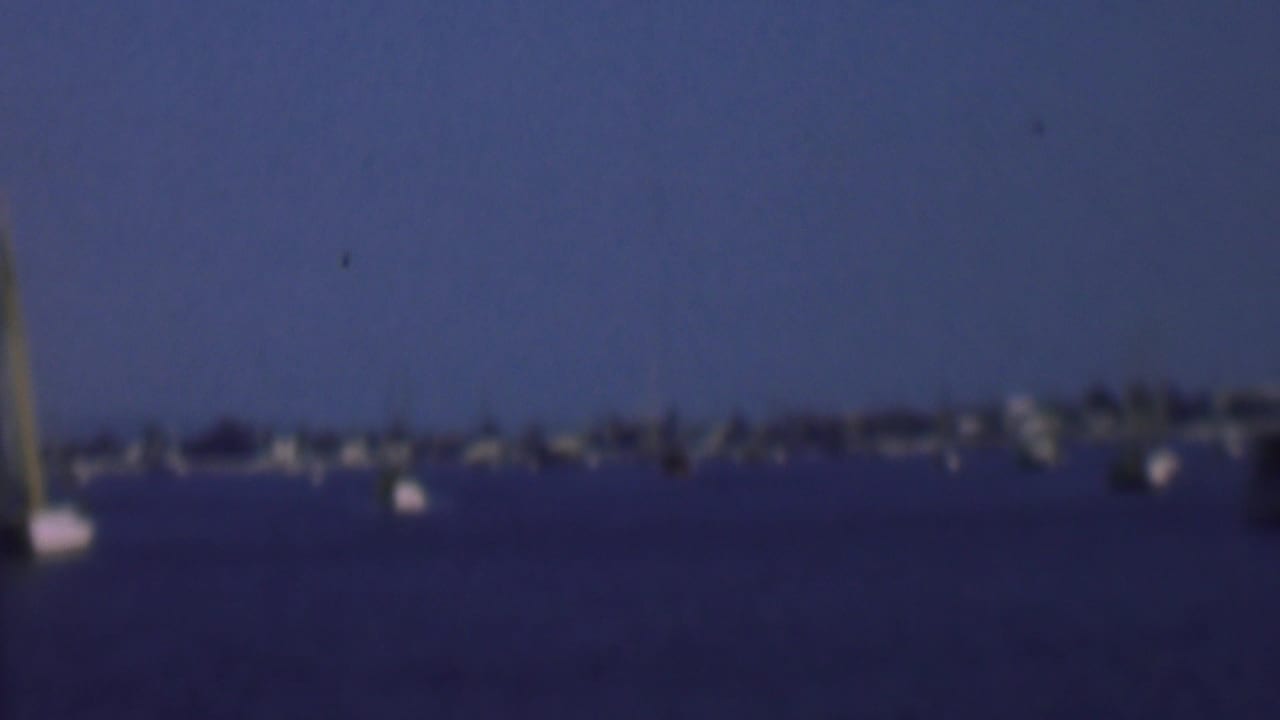 Soft 1961 8mm view across San Diego harbor, with small sailboats on blue water and a distant shoreline of low buildings and trees.
