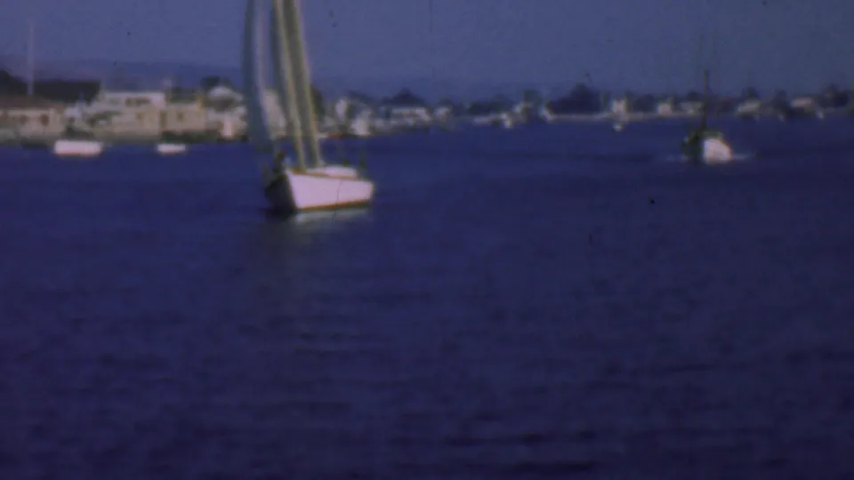 A sailboat with striped sails appears closer in the frame as the 1961 San Diego harbor shoreline remains soft in the background.