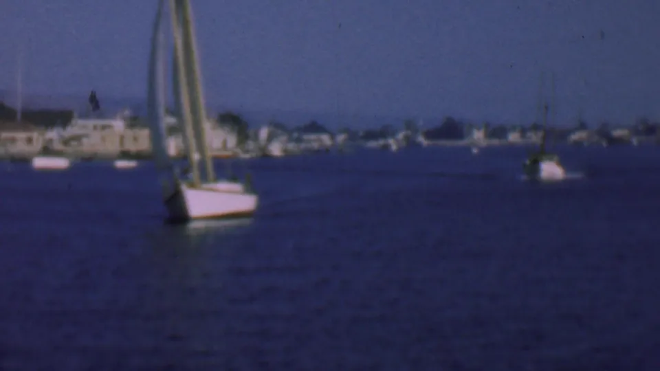 Closer 8mm view of a sailboat on San Diego harbor, with low waterfront structures, trees, and another small boat in the distance.