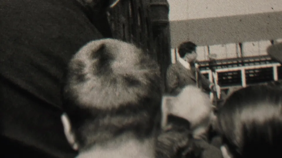 1937 Albany 8mm frame showing a crowded indoor meeting from behind the audience, with a suited man near a podium at the front of the room.