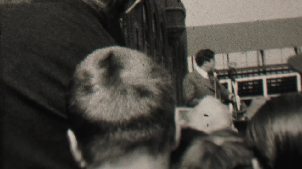 1937 Albany home-movie frame from inside a crowded meeting room, with foreground audience heads partly obscuring a suited speaker near the front.