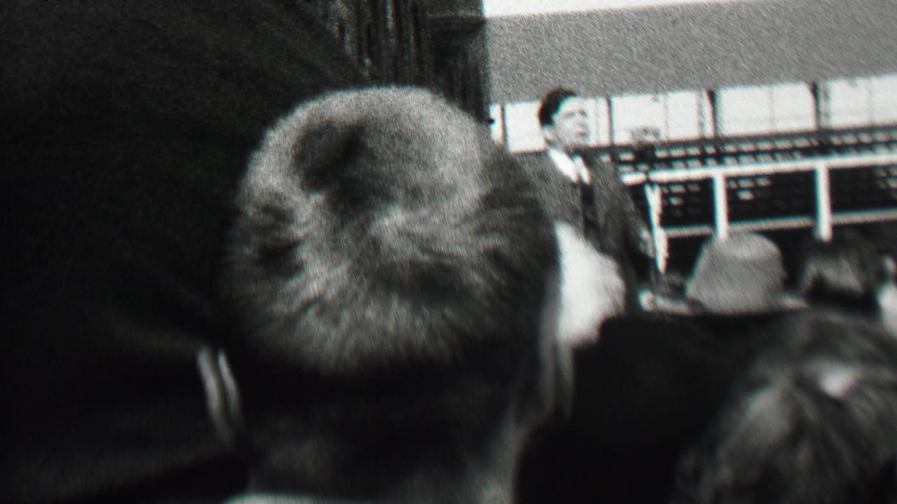 The speaker remains partly visible beyond the seated crowd, with the home-movie camera framed low from the audience area in the 1937 Albany gathering.