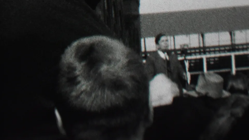 A seated audience, including a person in a brimmed hat, watches a suited man speaking near the front of an Albany public gathering in 1937.
