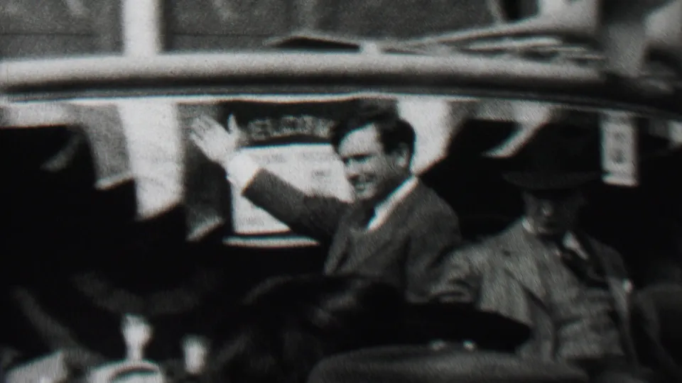 In Albany, New York, in 1937, a suited campaign figure appears to wave from an open car while men in hats sit nearby and spectators crowd the street.