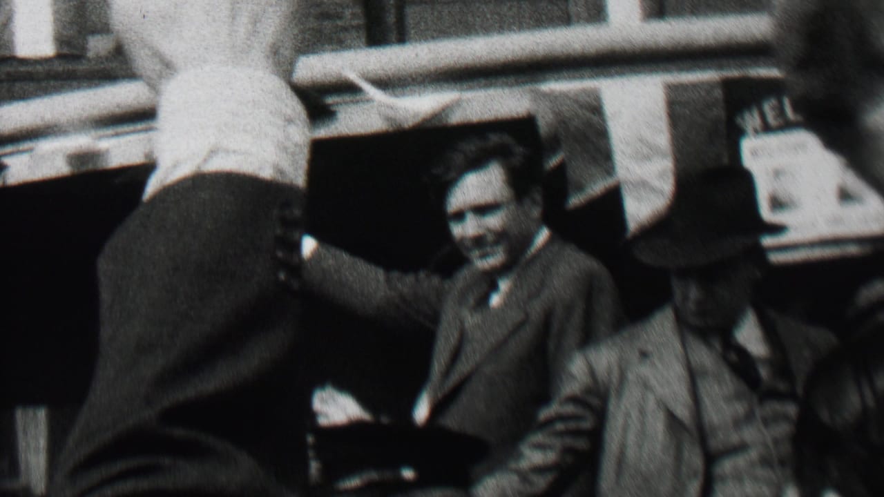 Waving suited man stands or leans from a moving car with passengers and storefronts behind him.