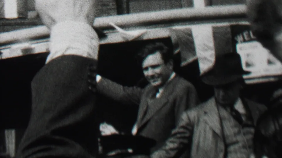 The 1937 Albany street scene shows the suited man smiling and waving from the moving campaign car, with hat-wearing passengers and building fronts behind him.