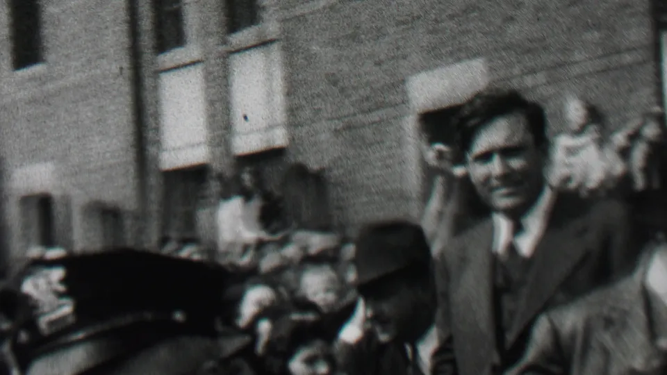 In Albany, New York in 1937, a suited man waves from within a dense public crowd as police officers and onlookers stand near a brick building entrance.