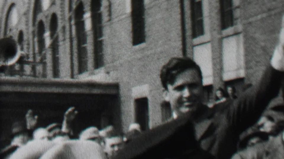 A suited man raises his arm in a close crowd in Albany in 1937, filmed from a handheld home-movie position near police and a brick building.