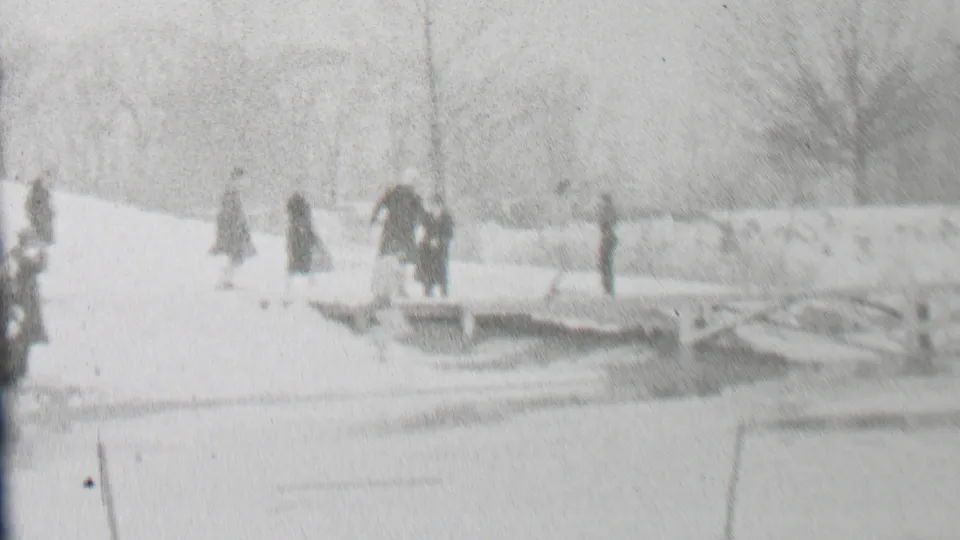 Several adults in winter clothing walk or hurry across a low wooden bridge in a snowy Albany park, seen through soft black-and-white 8mm exposure.