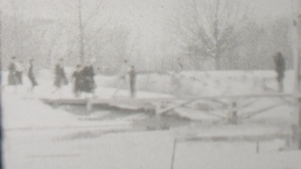 A small group stands or walks near the end of a wooden bridge in a snow-covered Albany park, with bare trees and open winter ground behind them.