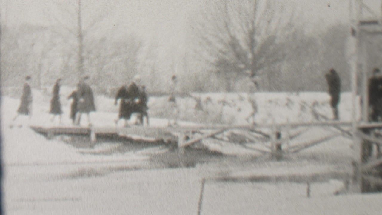 In 1937 Albany, New York, bundled pedestrians appear to cross a snow-covered bridge above a frozen or snow-covered waterway, with bare trees and bridge supports in the background.