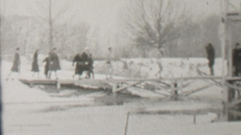 In 1937 Albany, New York, bundled pedestrians appear to cross a snow-covered bridge above a frozen or snow-covered waterway, with bare trees and bridge supports in the background.