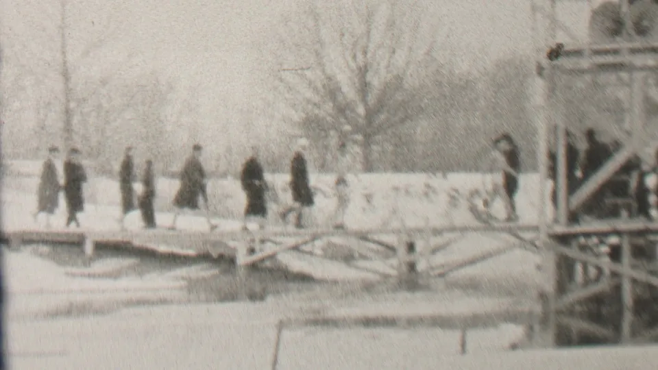 In 1937 Albany, New York, several pedestrians in dark winter clothing move across the snow-covered bridge, with bare trees and a metal or wooden framework visible to the right.