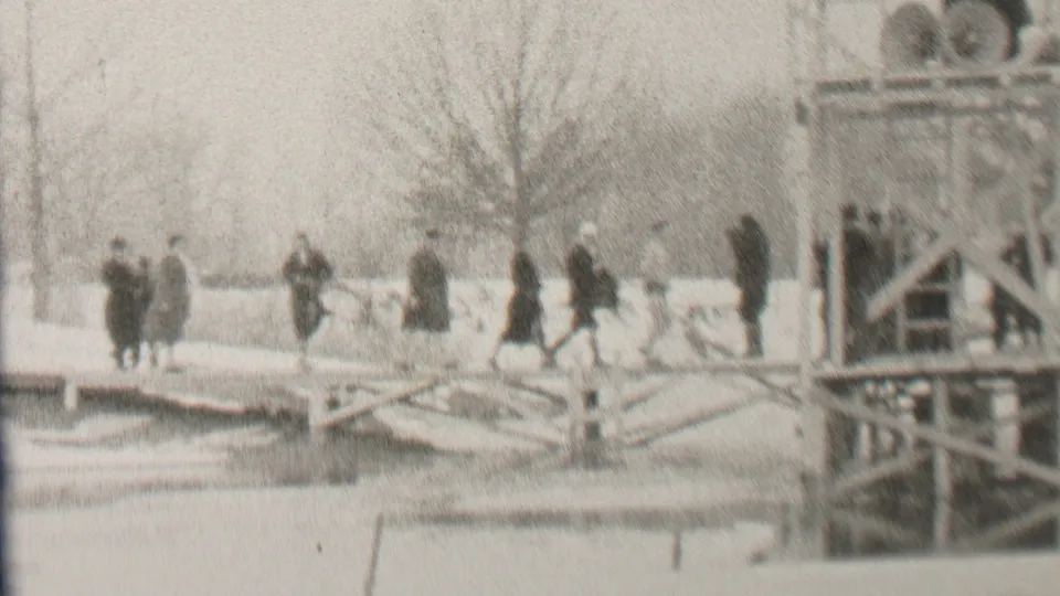 In 1937 Albany, New York, the home-movie view shows bundled people continuing across the snowy bridge, with a tall post, bare trees, and the bridge framework framing the scene.