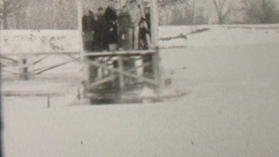 Grainy 8mm frame showing several bundled figures at a snow-covered wooden pier or platform, consistent with the Albany winter swimming scene described in the metadata.