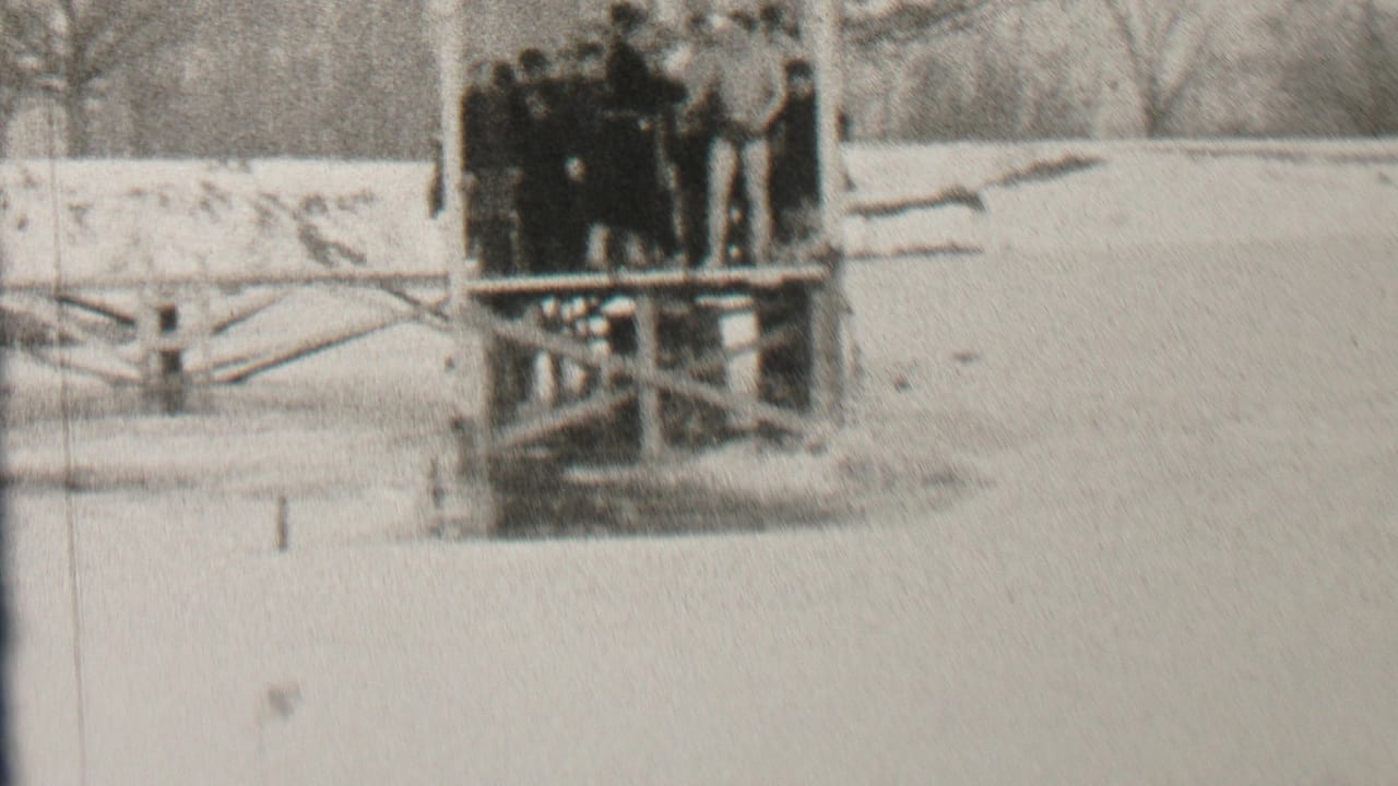 In supplied 1937 Albany home-movie footage, a group of young men in dark winter clothing gathers on a wooden waterside platform beside snow-covered ground and bare trees.