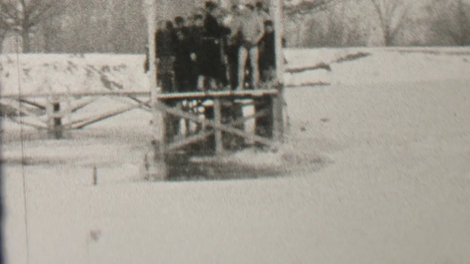 In supplied 1937 Albany home-movie footage, a group of young men in dark winter clothing gathers on a wooden waterside platform beside snow-covered ground and bare trees.