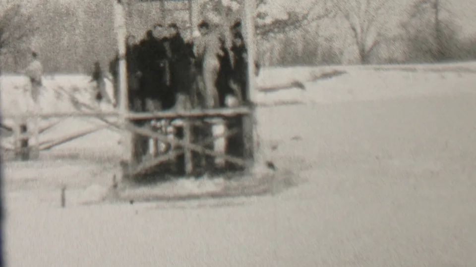Several young people in heavy coats stand together on the wooden pier, with the snowy ground and winter trees visible behind them in the Albany home-movie footage.