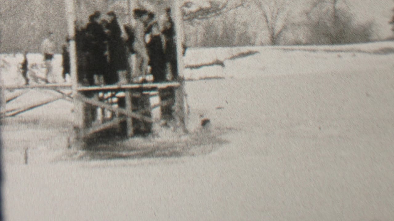 A group in winter coats stands close together on a wooden platform near snow.