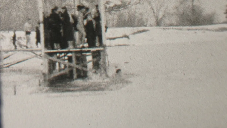 A cluster of people in dark winter clothing stands on a wooden platform in the snowy Albany winter scene, with open water visible below the structure.