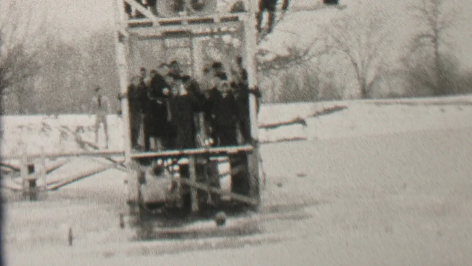 Albany, New York home-movie frame from 1937 showing men in winter coats grouped on a wooden lakeside platform with snow and bare trees behind them.