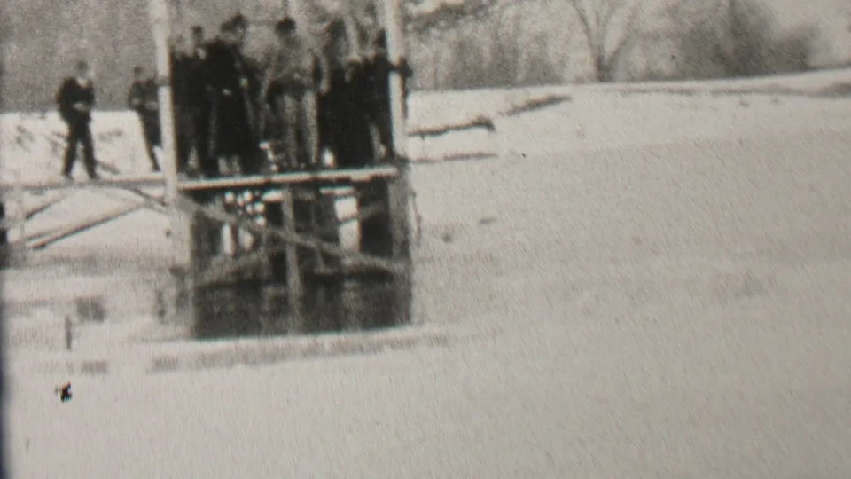 1937 Albany winter home-movie frame with men in dark coats gathered on a lakeside wooden platform and one figure standing to the side on snow.