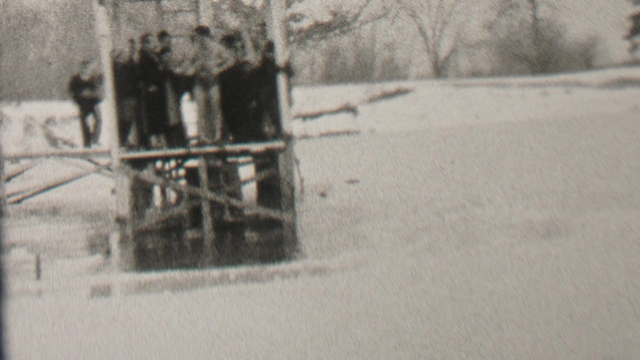 In the supplied 1937 Albany, New York home-movie context, a group in dark winter coats stands on a wooden dock over icy water with snow and bare trees nearby.