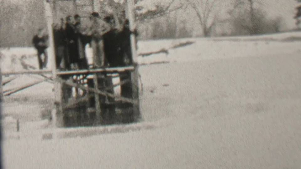 In the supplied 1937 Albany, New York home-movie context, a group in dark winter coats stands on a wooden dock over icy water with snow and bare trees nearby.