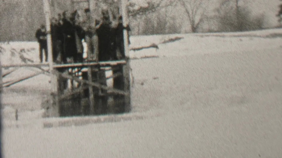 The frame shows the Albany winter scene with several people clustered on a dock above icy water, framed by a pale snowy bank and leafless trees.
