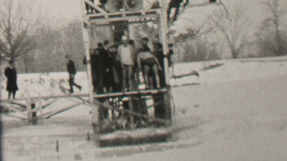 In 1937 Albany, New York, bundled spectators gather around a wooden platform on a frozen or icy body of water, with snow and bare trees in the background.