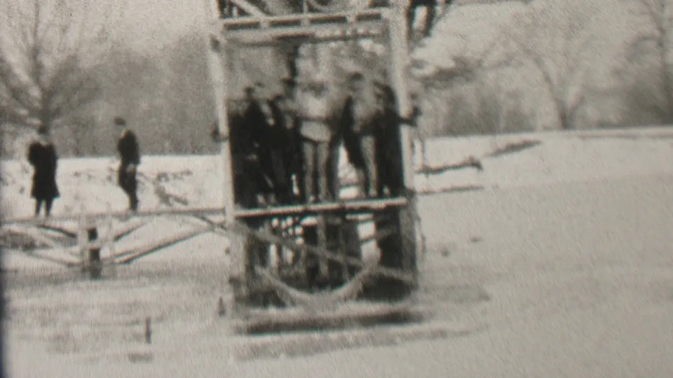 Spectators in heavy winter clothing stand on and near a wooden platform over icy water in Albany, with a snowy bank and leafless trees beyond.