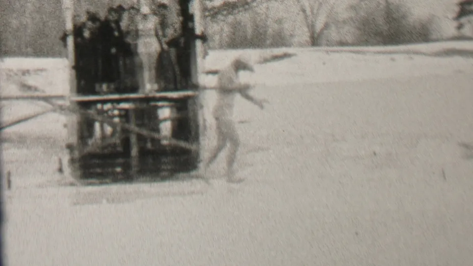 In 1937 Albany, New York home-movie footage, a swimsuit-clad man moves across ice beside a wooden platform where bundled spectators stand.