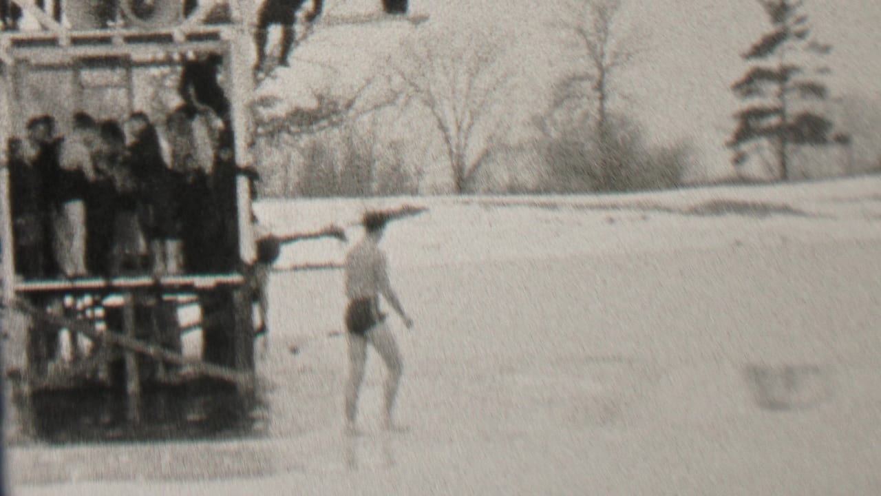 A man in a swimsuit stands on a frozen surface near a wooden dock with spectators.
